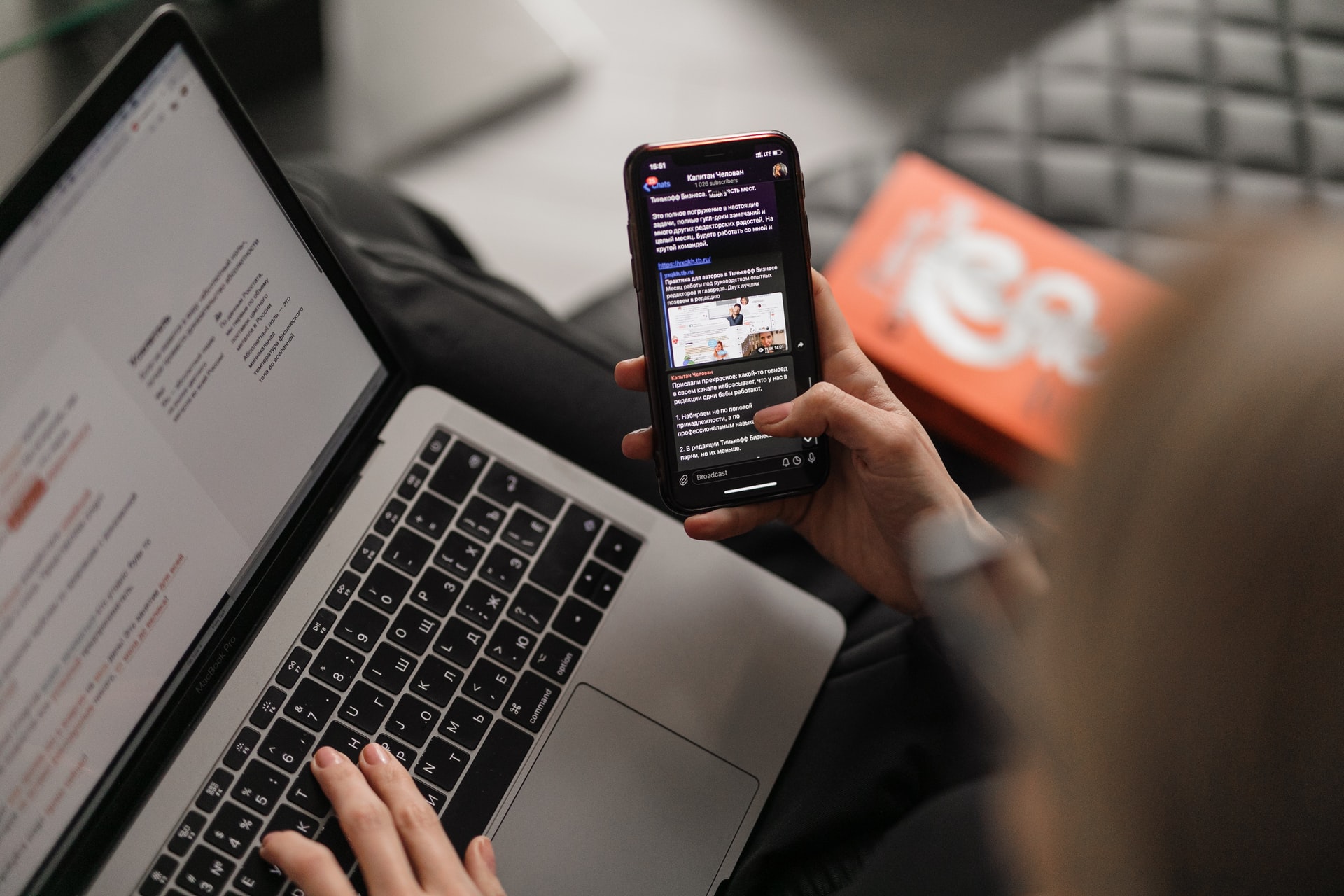 Obrazek przedstawiający laptopa oraz smartfona, z którego korzysta kobieta. Widać jej dłonie i część włosów.